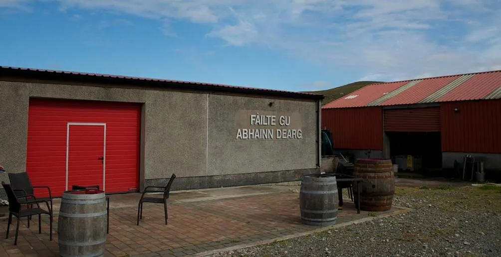 Einige Fässer und Stühle vor dem Lagerhaus Abhainn Dearg mit einer leuchtend roten Tür an einem schönen Tag