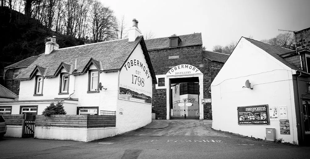 A black and white photo of Tobermory's distillery building made of dark stone located behind two houses