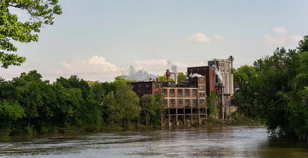 Blanton's distillery standing by a river surrounded by green trees with steam coiling out