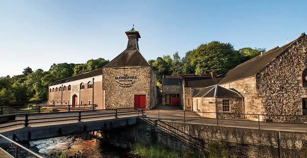 The distillery building made of brown bricks with red doors and trees in the background viewed from the other side of the spring