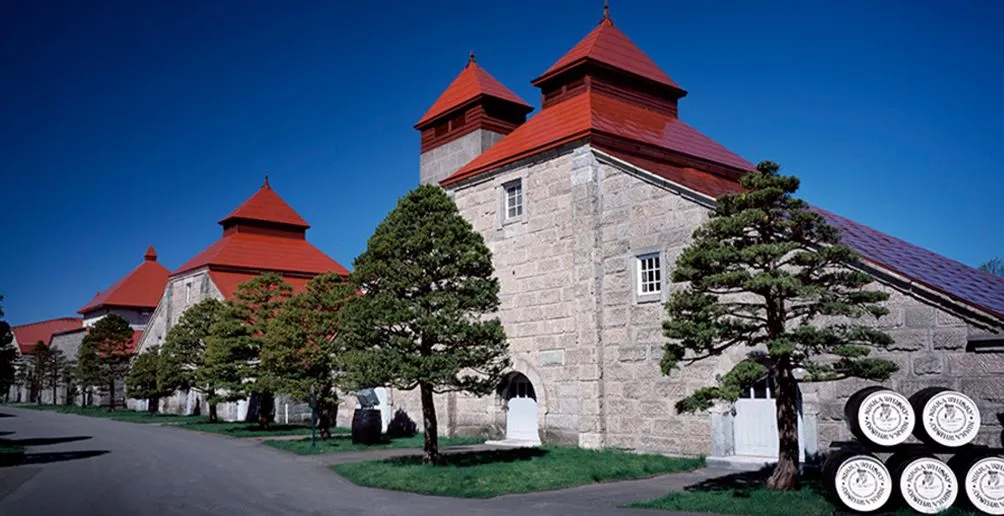 Yoichi distillery's grey stone buildings with pagoda-style red rooftops hidden behind trees on a sunny day