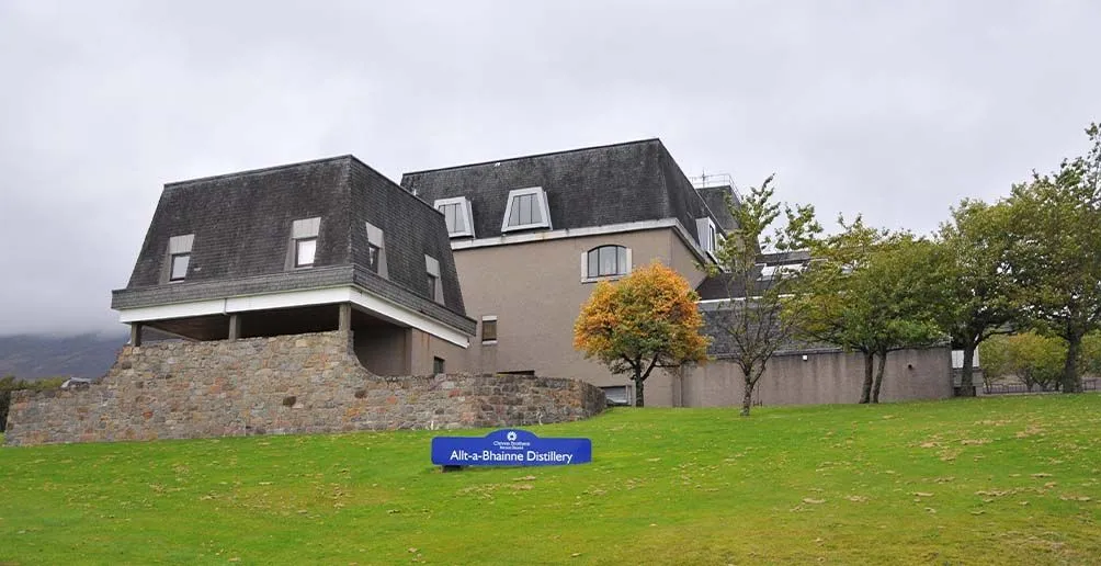 Alt-a-Bhainne distillery with dark roofs and beige walls with its name sign lying on the grass in the front