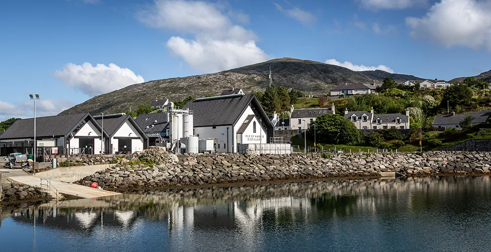 Harris distillery with white walls and dark roof located at the foot of a mountain next to the stream of Abhainn Cnoc a ’Charrain