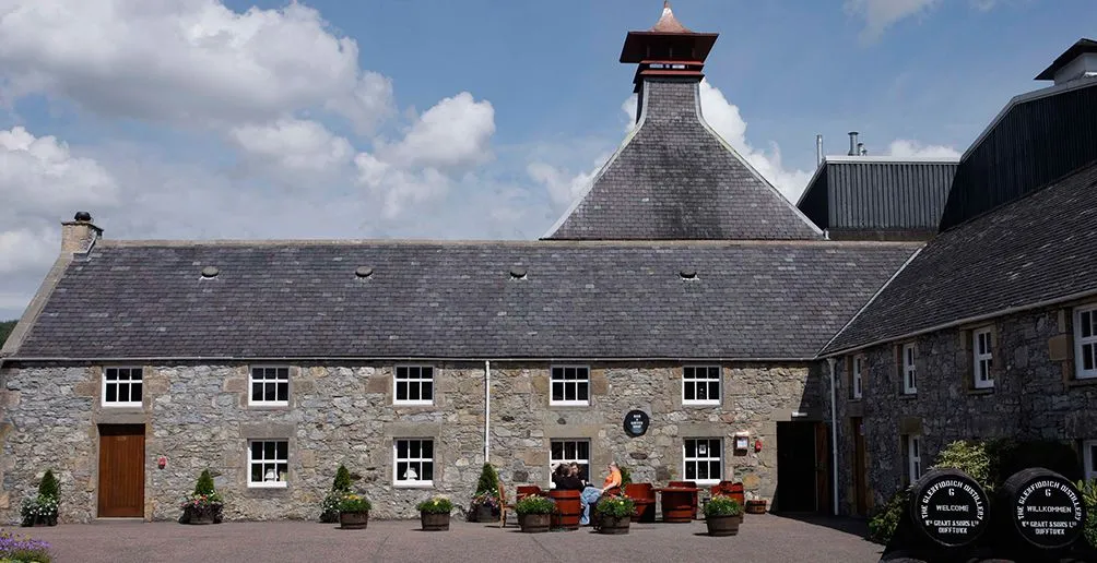 Visitors standing in front of Glenfiddich's distillerly stone building with grey padoga style roof on a sunny day