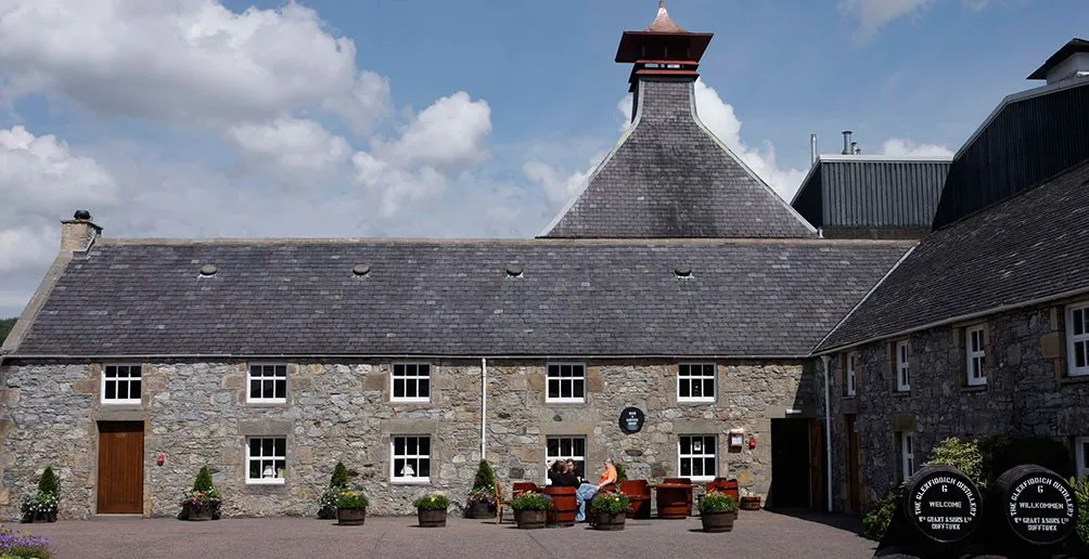 Besucher stehen an einem sonnigen Tag vor dem steinernen Destilleriegebäude von Glenfiddich mit dem grauen Dach im Padoga-Stil