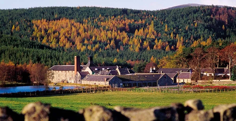 Royal Lochnagar distillery located next to a lake at the foot of a mountain with trees starting to turn yellow in early autumn
