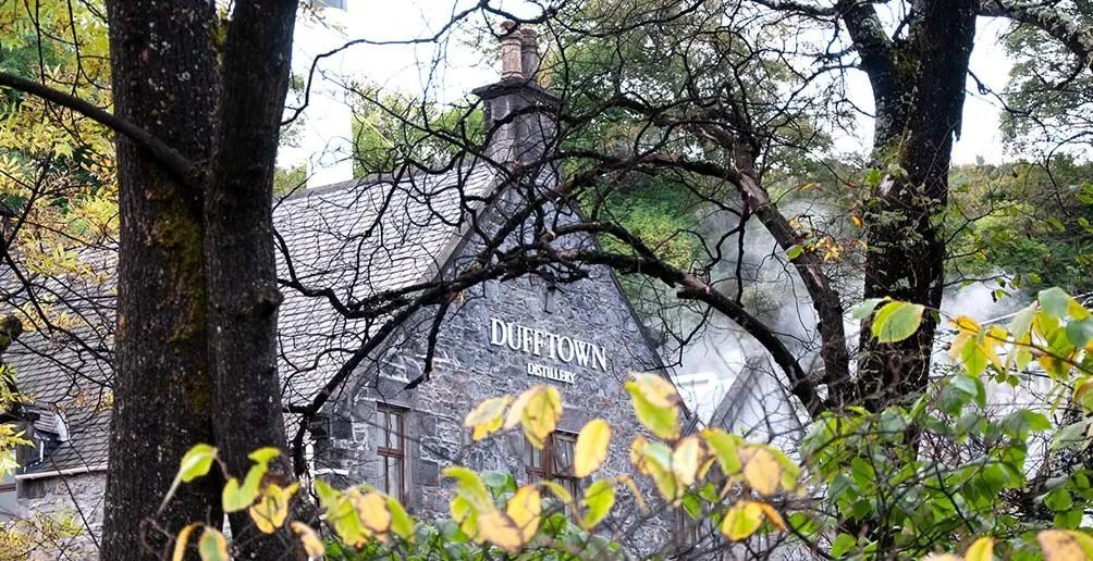 Dufftown distillery made of stone with steam coiling out of the chimney hiding itself among nature