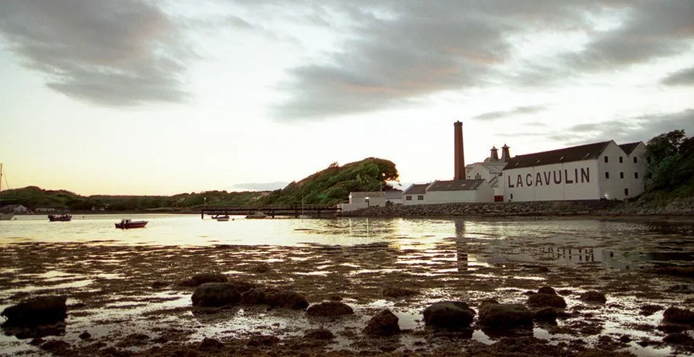 Lagavulin distillery's white buildings with its name painted on a side wall viewed from the river Abhainn nam Beitheachan during a low tide