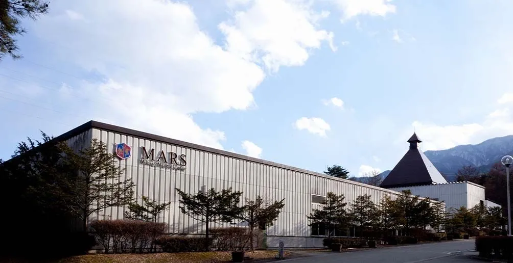 Mars Shinshu's warehouse with the company's name and logo on the wall hiding behind trees and bushes on a nice day
