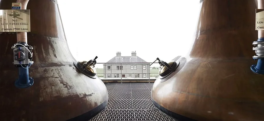Front side of a distillery building standing in the middle viewed from behind two copper pot stills on two edges of the picture