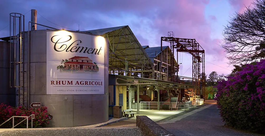 Clement distillery with its name and logo attached on the large tank in the front surrounded by nature on a blue sky background