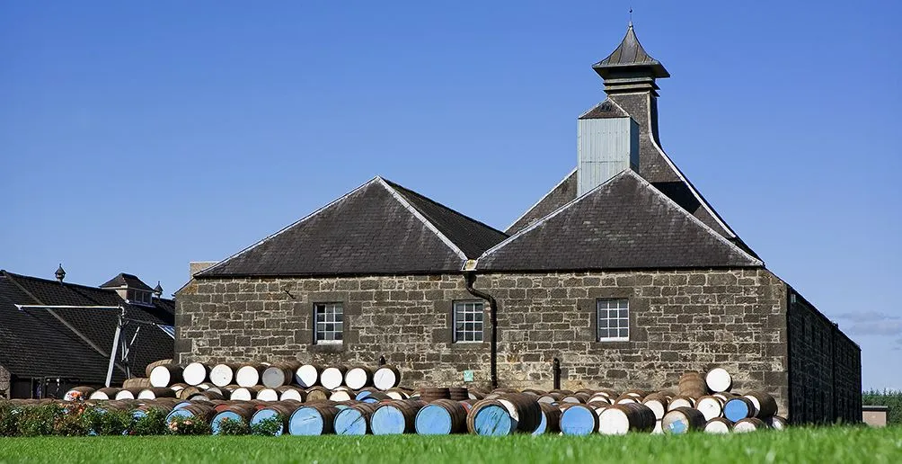 Casks with blue and white lids lying in front of Benriach's distillery building with blue sky in the back ground