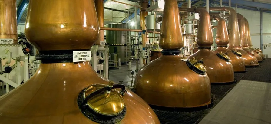 Eine Reihe von sieben kupfernen Pot Stills im Inneren des Destillierhauses mit Maischebottichen im Hintergrund