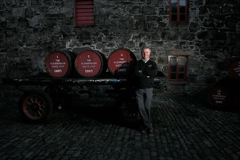 Glendronach's employee leaning against a cart carrying three red-lid casks in front of Glendronach's warehouse made of stone