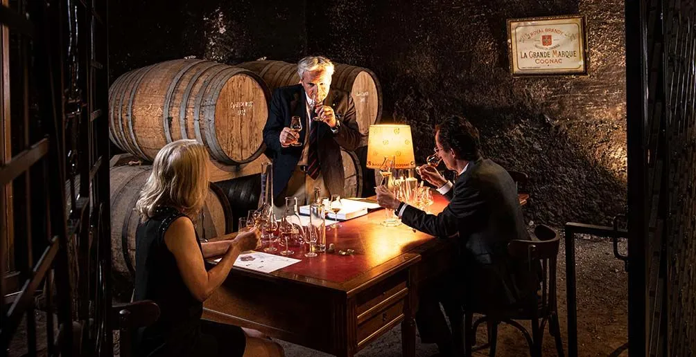 Three connoisseurs tasting Camus cognac in front of some casks stacked on shelf