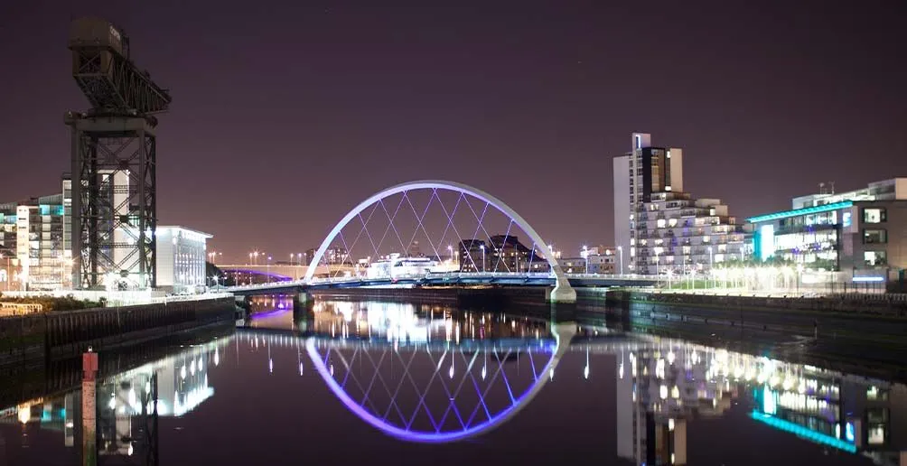 Squinty bridge with lights on reflecting itself in the river Clyde with Finnieston crane on the left bank and illuminated building on the right
