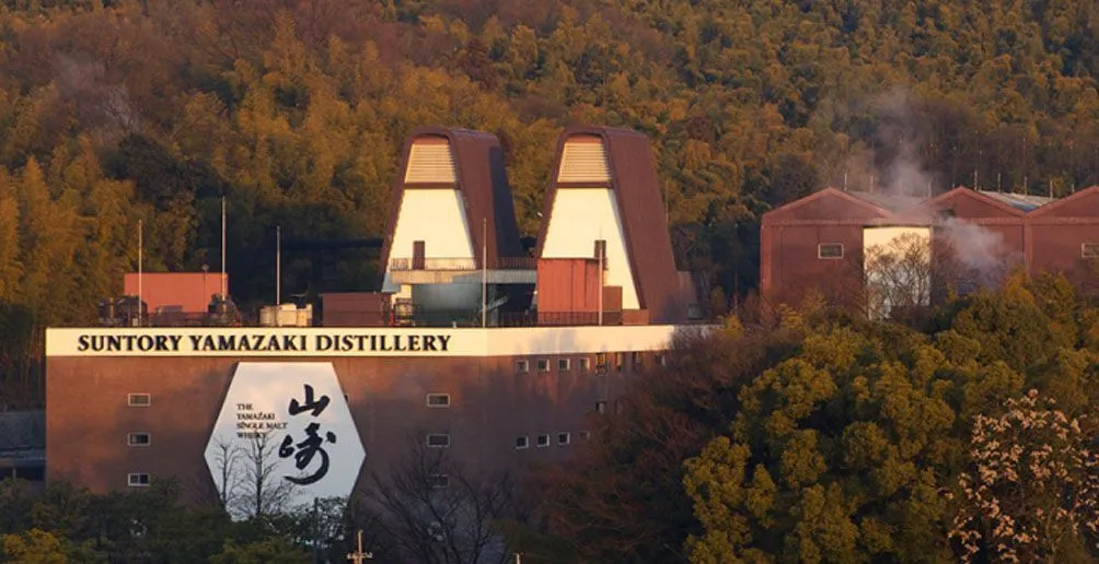 Das Gebäude der Yamazaki-Destillerie mit roten Wänden und Dächern, mit dem Namen und dem Logo an der Fassade, umgeben von Natur