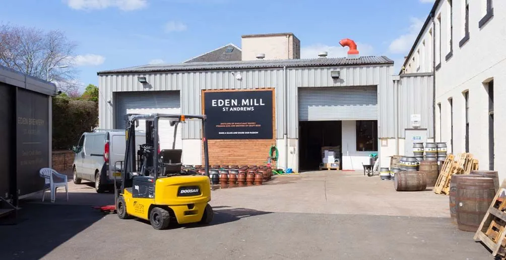 Van, forklift vehicle and casks standing outside of Eden Mill's warehouse with its logo on the wall
