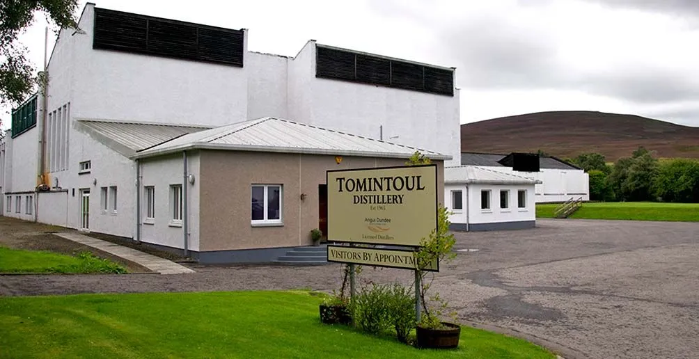 Tomintoul's white distillery building behind its yellow welcome board with a bald hill in the background