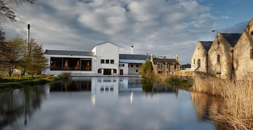 Exterior picutre of Royal Brackla distillery with its white still house in the middle and brick warehouse on the right viewed from the lake