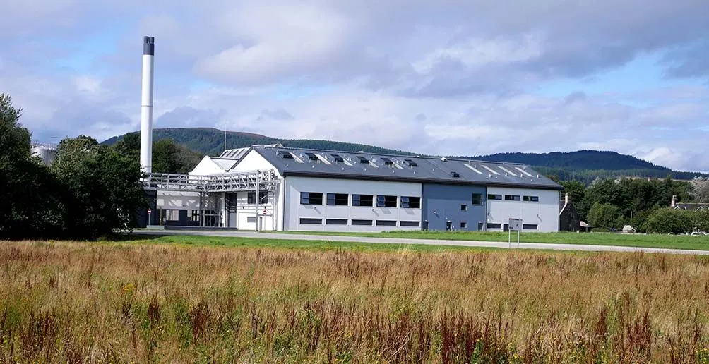 Mordern white building of Teaninich distillery surrounded by green trees and grass fields with a hint of rolling mountains in the background