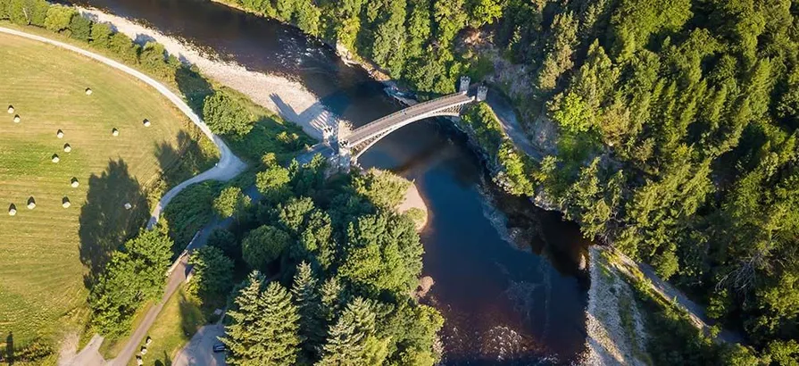 A picture of Spey River running in between of a wood and a grass field under a bridge taken from above