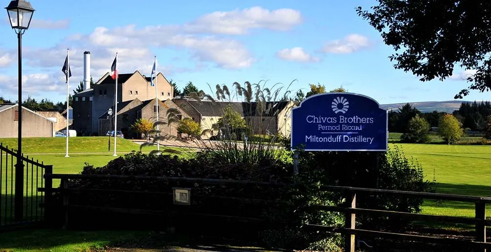 Miltonduff distillery appearing behind its blue direction sign on a sunny day