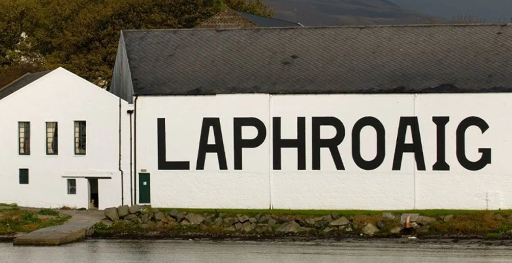 Laphroaig distillery's white building with its name painted on the wall located at the foot of a mountain viewed from the river
