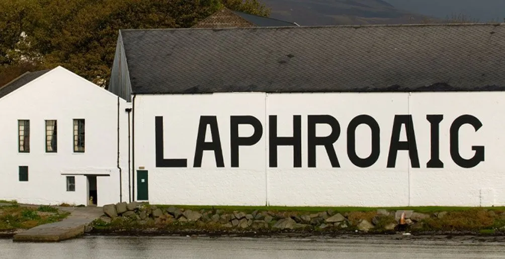 Das weiße Gebäude der Laphroaig-Brennerei mit ihrem Namen an der Wand am Fuße eines Berges vom Fluss aus gesehen