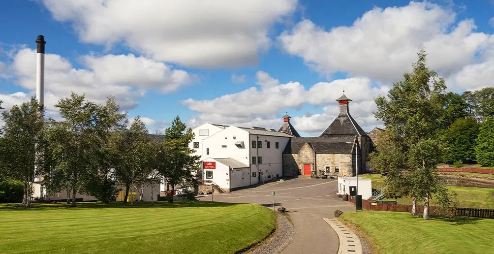 Distillery buildings of Cardhu surrounded by green trees with blue sky and white clouds in the background