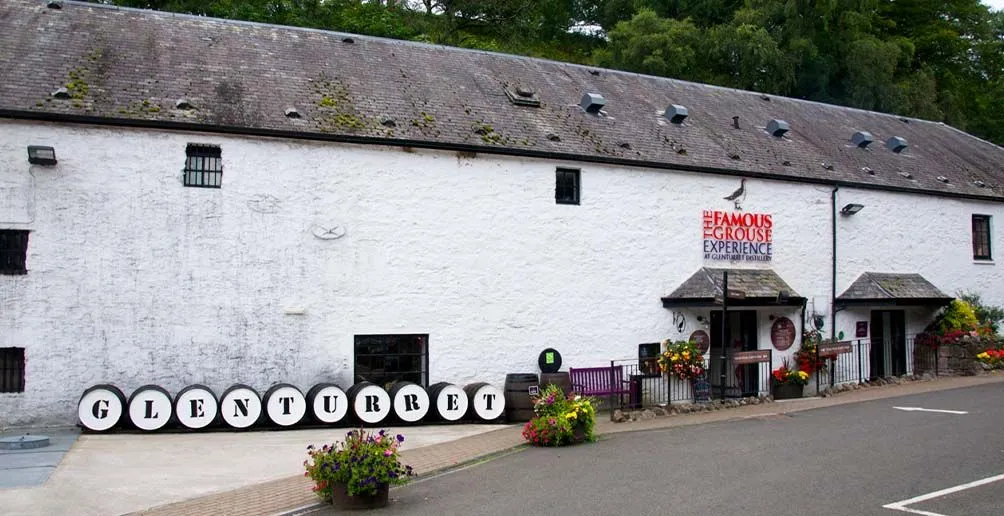 Exterior of Glenturret's white distillery building decorated with casks with the distillery's name on the lids and flowers in full bloom