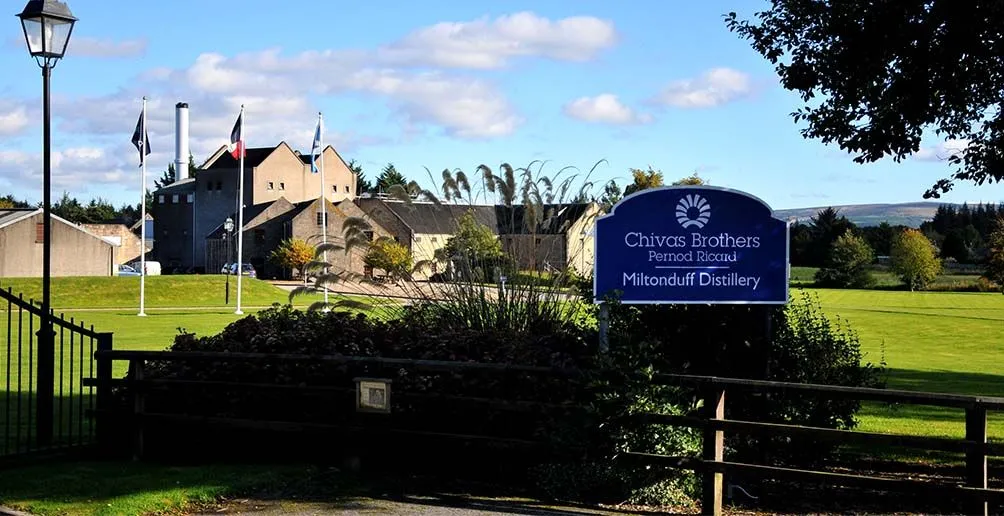 Miltonduff distillery located on a green yard with mountains and blue sky in the background viewed from its welcome sign