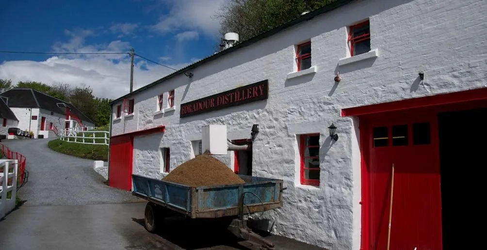 A cart full of barley standing in fron of Edradour distillery with white walls and bright red doors and windows