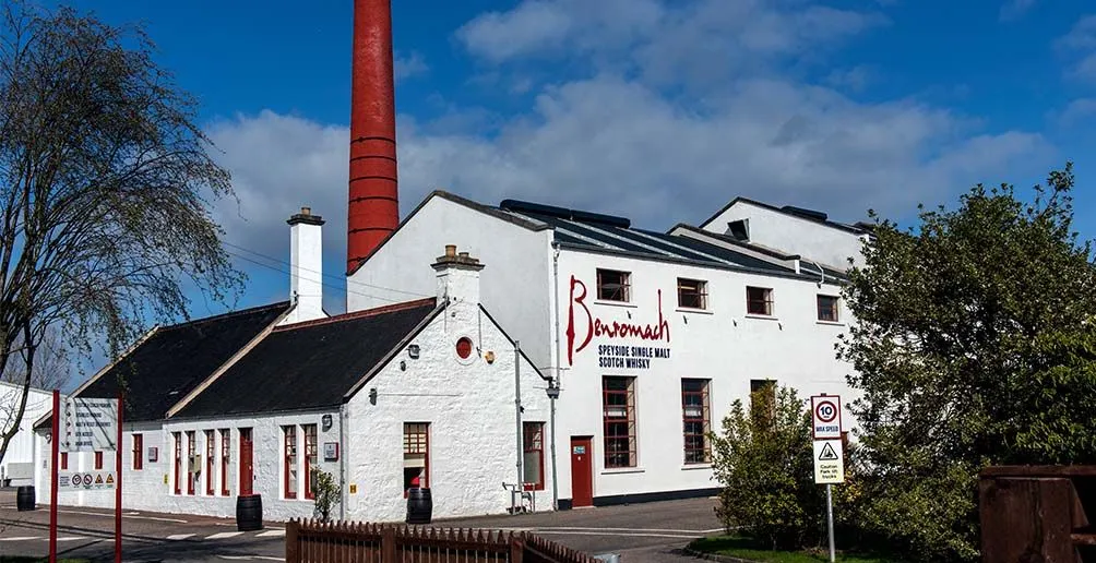 Benromach's white distillery building with its name painted in red on the wall and a red chimney standing behind