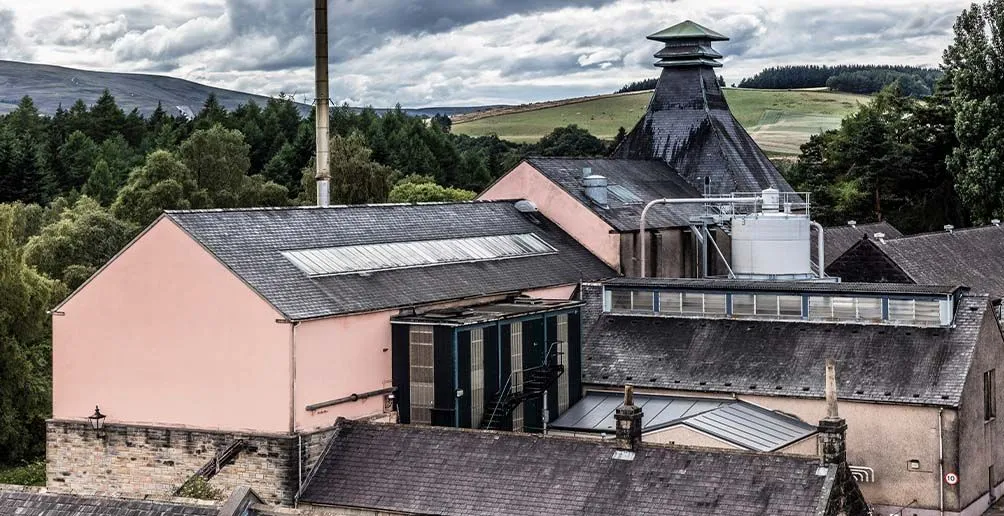 Knockando distillery buildings with pink walls and dark roofs located among nature with rolling hills in the background