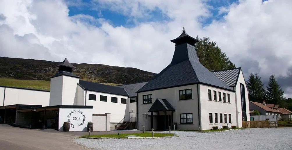 Entrance to Ardnamurchan distillery with white walls and dark roofs located at the foot of a hill