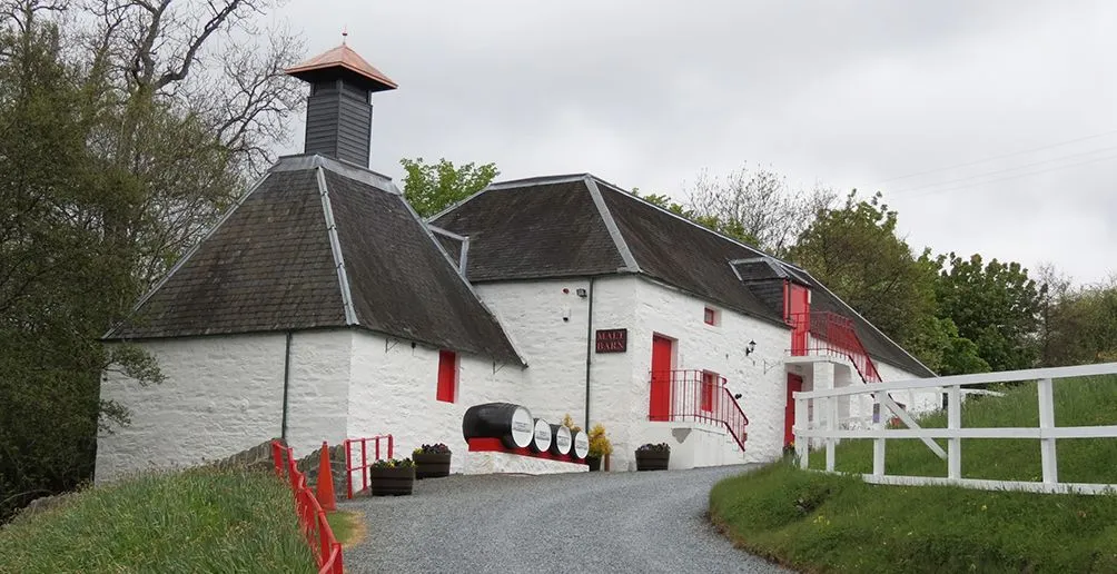 Path to Edradour's distillery building with its white house facade and red doors surrounded by green trees on a cloudy day