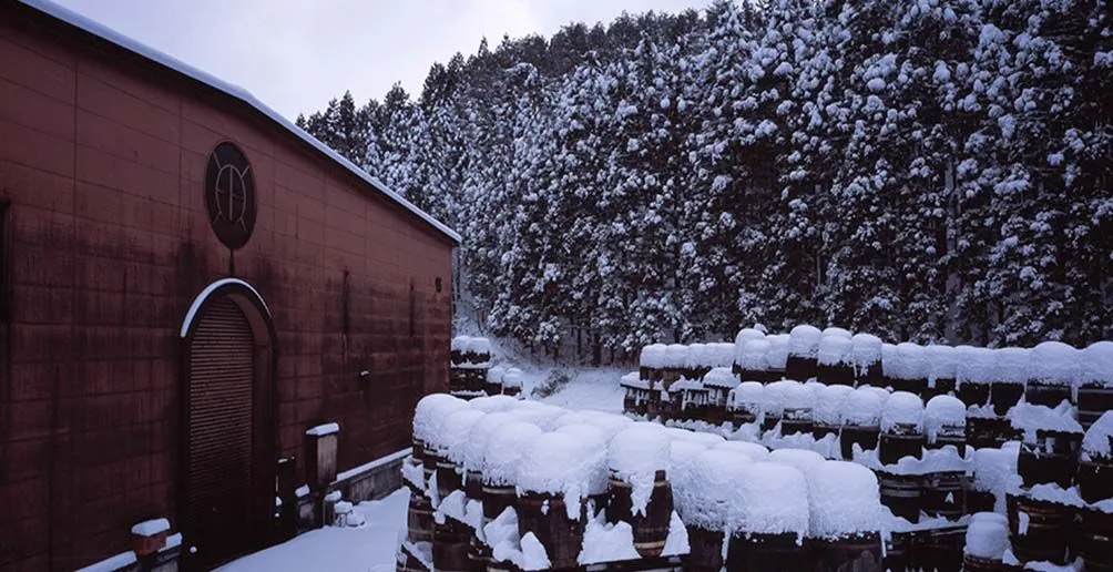 Gestapelte Fässer vor dem roten Lagerhaus von Miyagikyo, das im Winter mit Schnee bedeckt ist