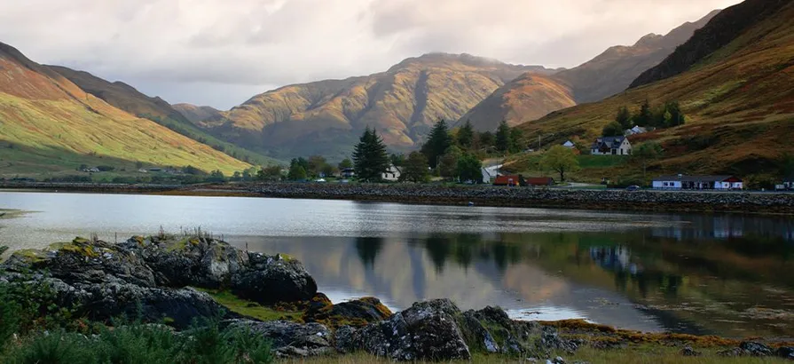 A small town at the foot of a mountain in the east Highlands surrounded by green pine trees viewed from the other bank of the lake