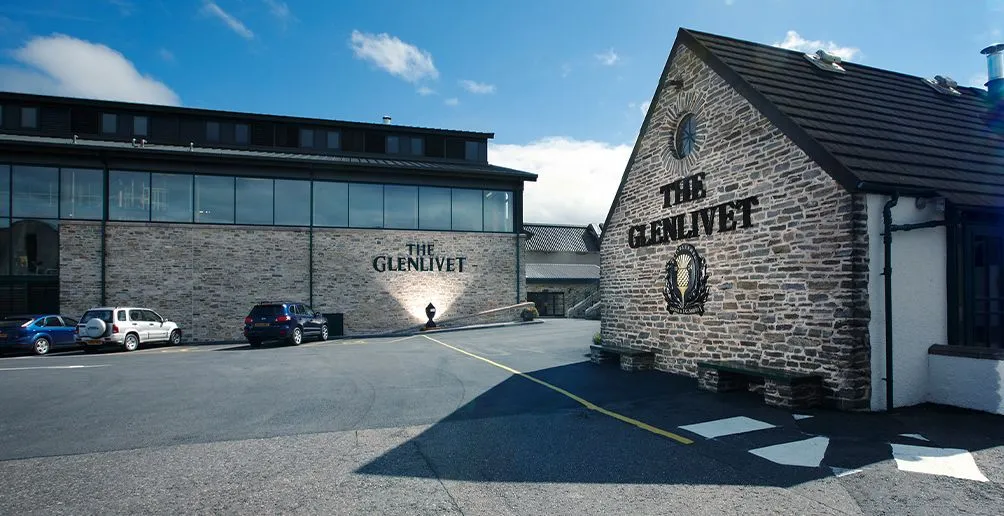 Parkplatz vor den Gebäuden der Glenlivet-Destillerie mit dem beleuchteten Namen und Logo an der Stirnwand