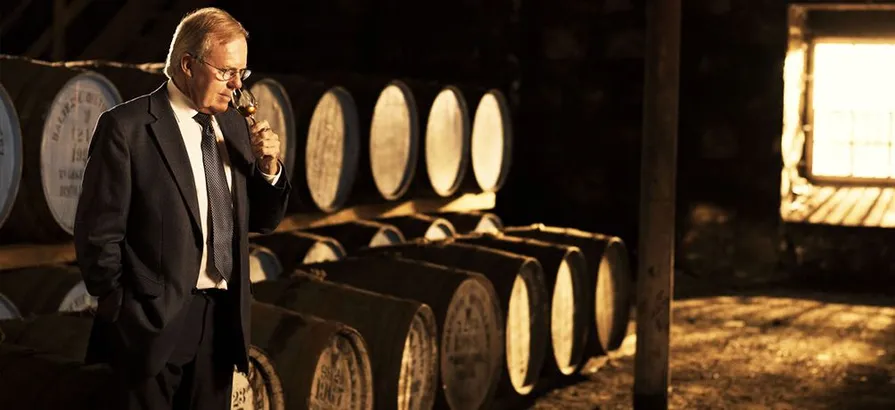 Malt Master David C. Stewart with a nosing glas in hand standing in front of rows of casks in Balvenie warehouse