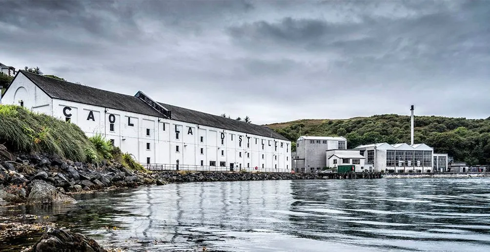 Caol Ila distillery's white buildings with its name painted on the wall located at the foot of a green hill viewed from the shore