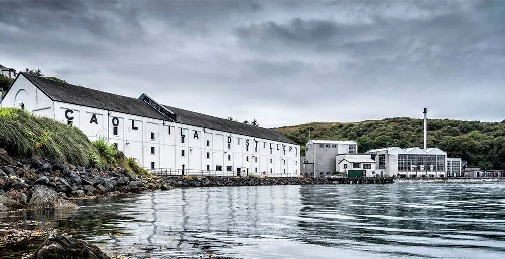 Die weißen Gebäude der Caol Ila Destillerie mit ihrem Namen an der Wand am Fuße eines grünen Hügels von der Küste aus gesehen