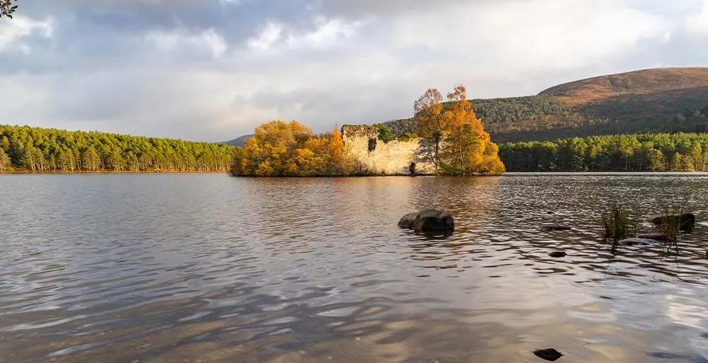 The beautiful Spey river surrounded by green trees on both banks with the footage of an ancient building on an island in the middle and rolling hills in the background