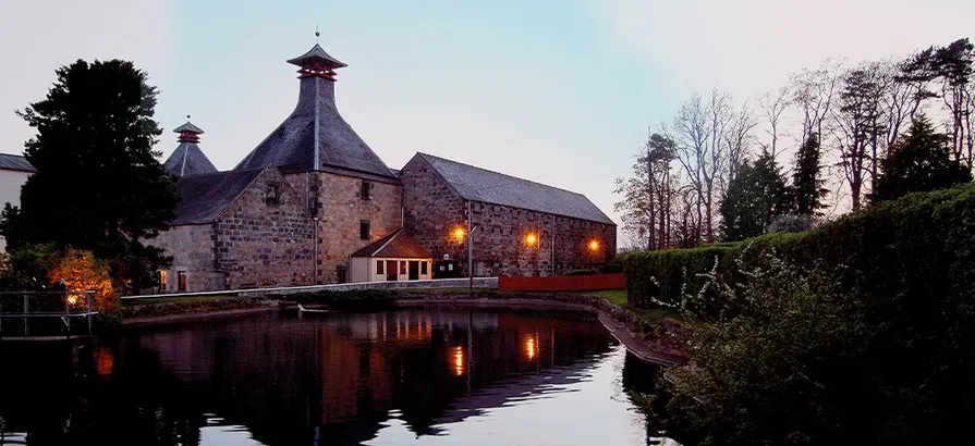 Cardhu distillery's brick-building illuminated in twilight with a lake in the front yard