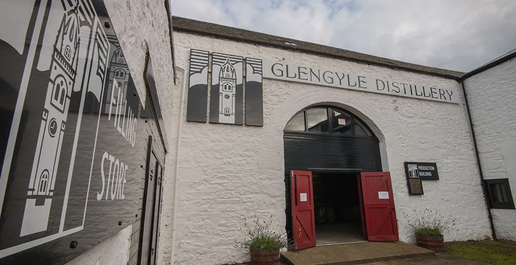 Entrance to white production building of Glengyle distillery with open red doors and its brand name and logo attached on the walls